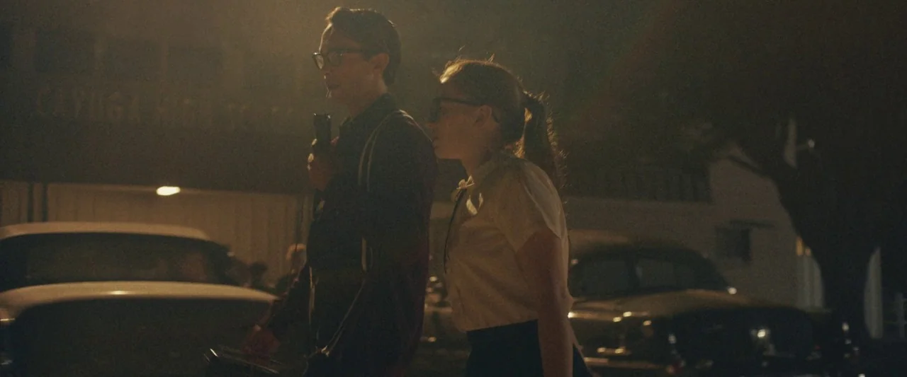 The Vast of Night (2019) movie still: medium — A night scene in a 1950s parking lot with vintage cars parked nearby. A teenage boy and girl stand…