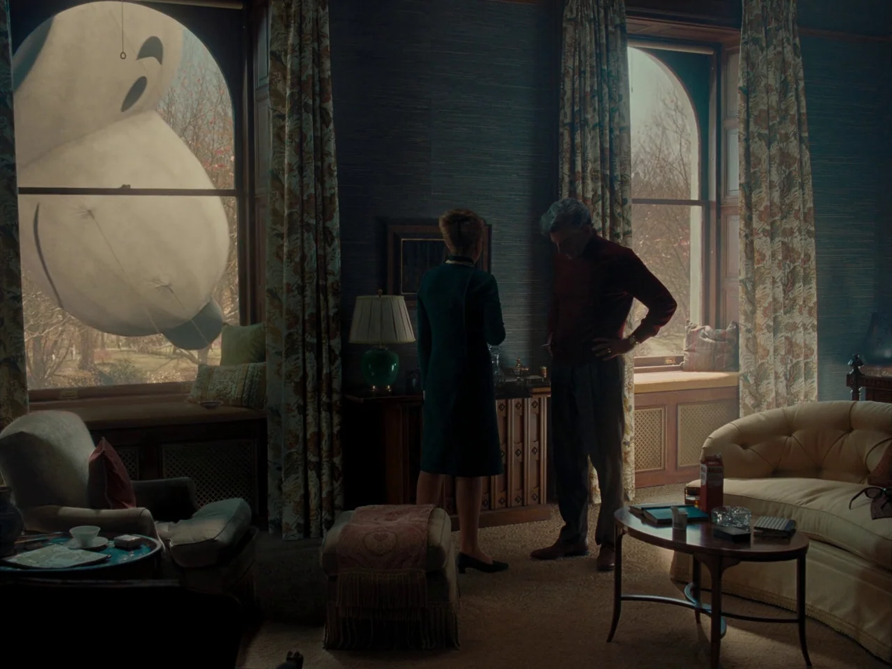 Maestro (2023) movie still: wide — A mid-century living room features patterned curtains, textured dark blue wallpaper, and wood furniture. A man…