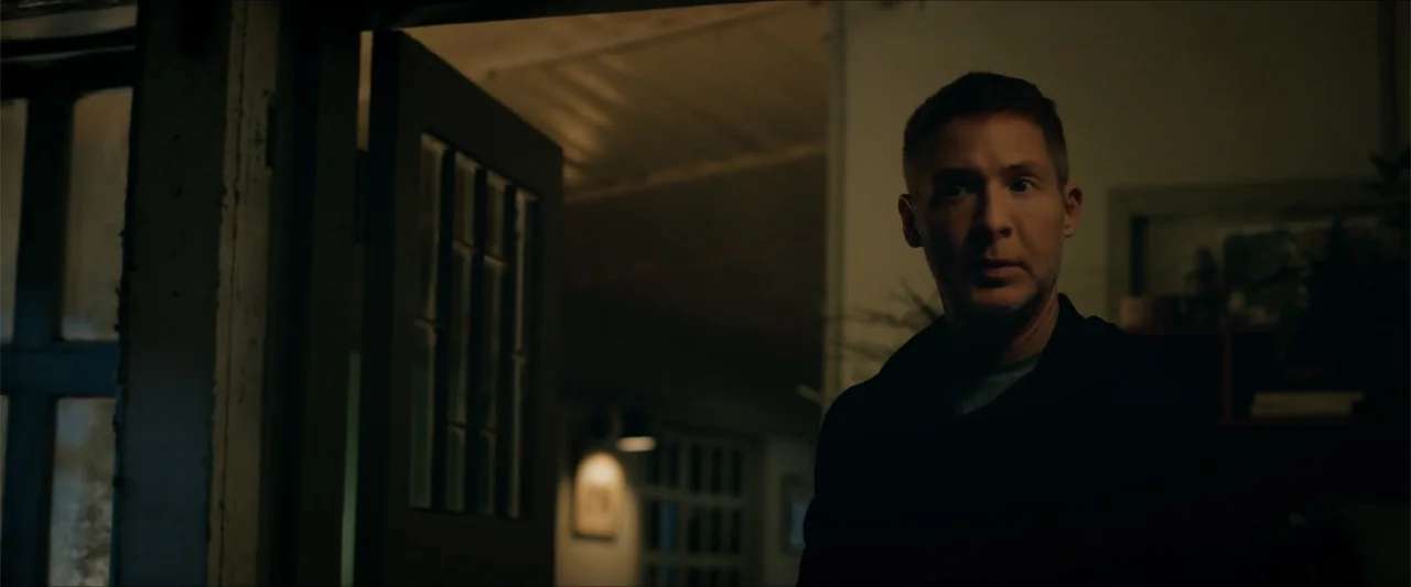 Checkmate (2015) movie still: medium — A dimly lit residential room features a dark open door leading into shadows. A man with short hair and a…