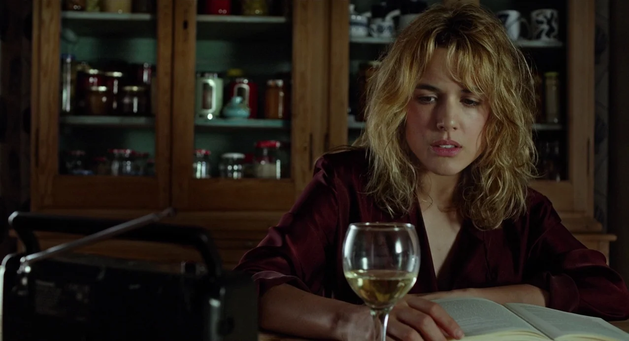 Julieta (2016) movie still: medium — A wooden table is set in a dimly lit room with a cabinet of jars. A woman sits reading a book next to a glass…