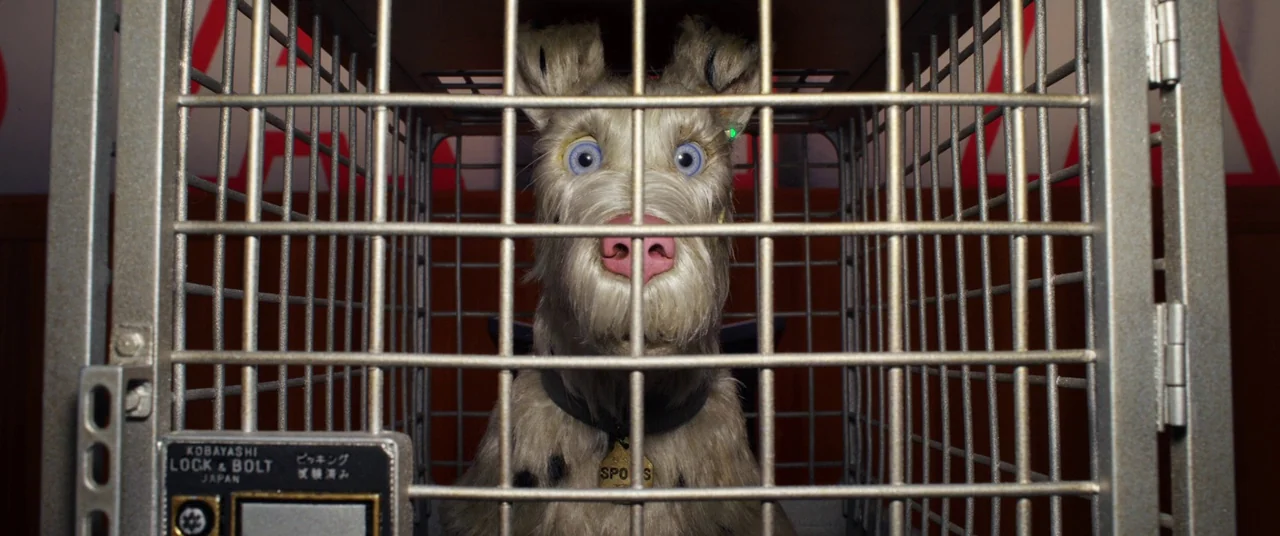 Isle of Dogs (2018) movie still: closeup — A stop-motion dog sits inside a grey metal transport cage with a latch. The animal looks directly out…
