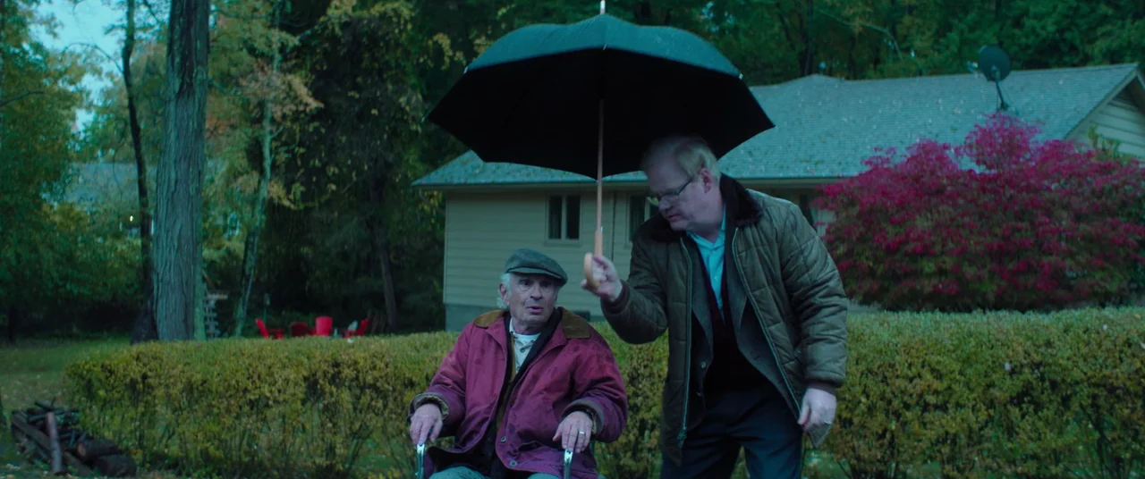 Linoleum (2022) movie still: medium — A residential yard features a trimmed green hedge and a suburban house. An adult man holds a black umbrella…