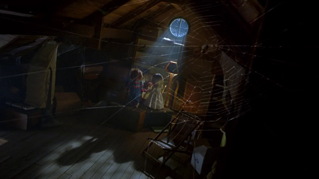 Seed of Chucky (2004) movie still: wide — A dark attic is filled with stored boxes, an old chair, and a spiderweb. Two small dolls sit on a trunk in…
