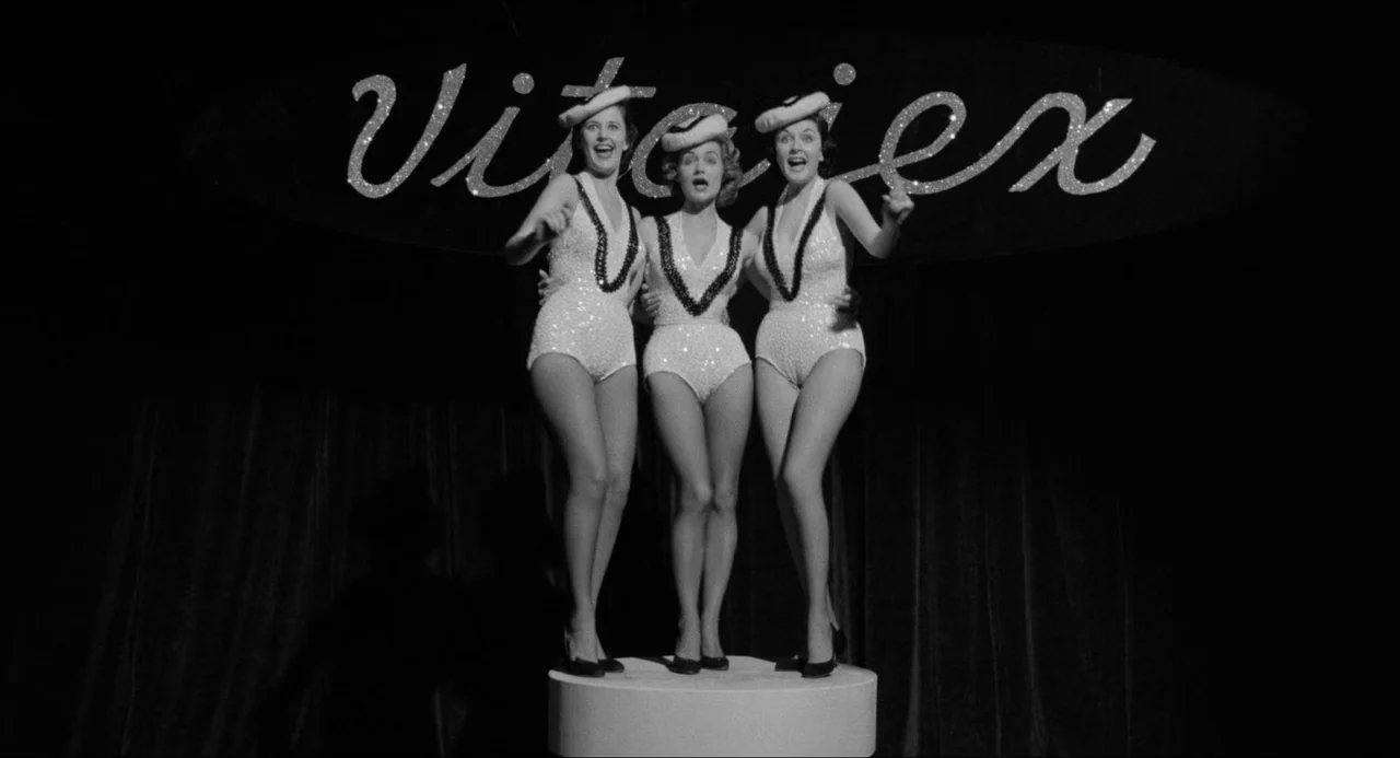 A Face In The Crowd (1957) movie still: fullbody — Three women in sequined costumes stand on a cylindrical platform in a darkened venue. They smile…