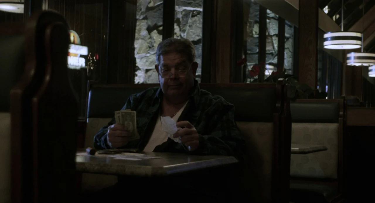 Happer’s Comet (2022) movie still: medium — A dimly lit restaurant booth with dark green vinyl upholstery and stone wall backing. An older man sits…