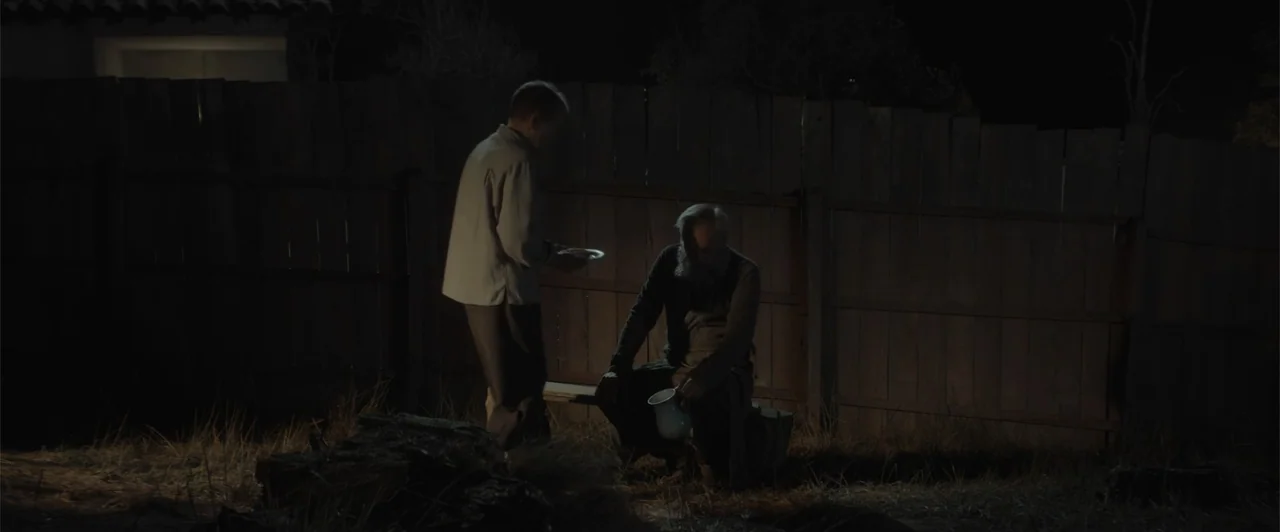 My Neighbor Adolf (2022) movie still: medium — A dark outdoor space enclosed by a tall wooden fence at night. An older man sits on a wooden bench…