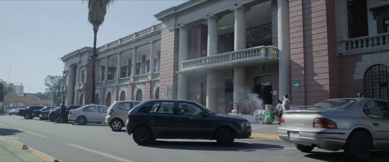 Radical (2023) movie still: wide — A historic brick building with white classical columns stands along a paved city street. Several cars are parked…