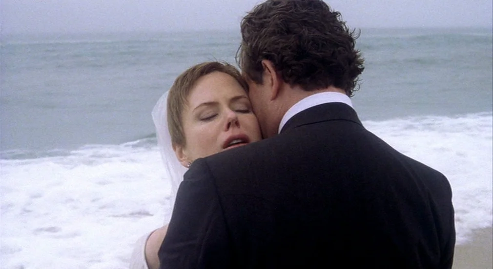 Birth (2004) movie still: medium — An overcast beach setting with gentle waves breaking on the sandy shore. A man in a dark suit embraces a woman in…