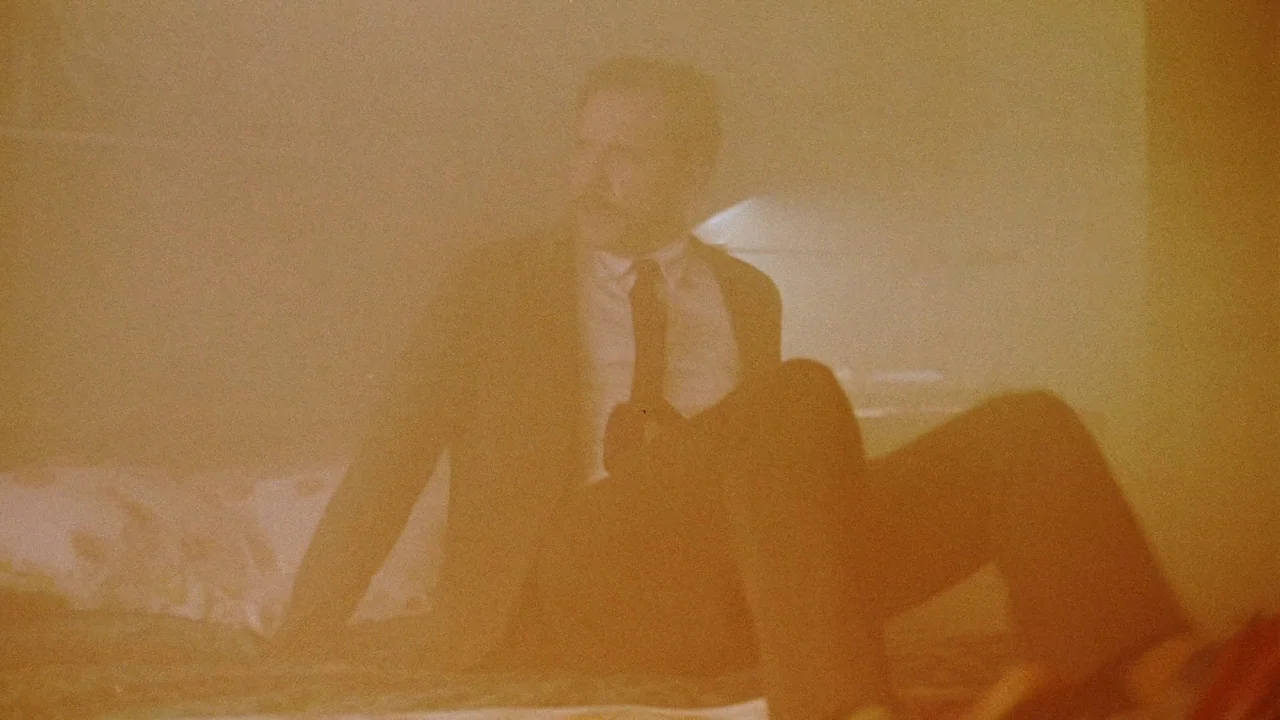 Strawberry Mansion (2021) movie still: medium — A man in a dress shirt sits on a bed in a dim, orange-hued room. The air appears hazy or filled with…