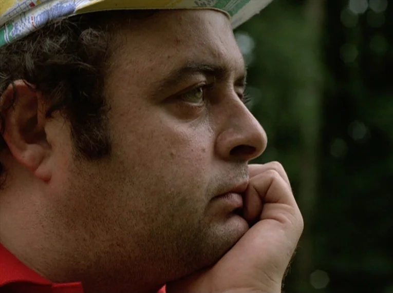 Keep Your Right Up (1987) movie still: closeup — A man wearing a cycling cap rests his chin on his hand. The background is composed of out-of-focus…