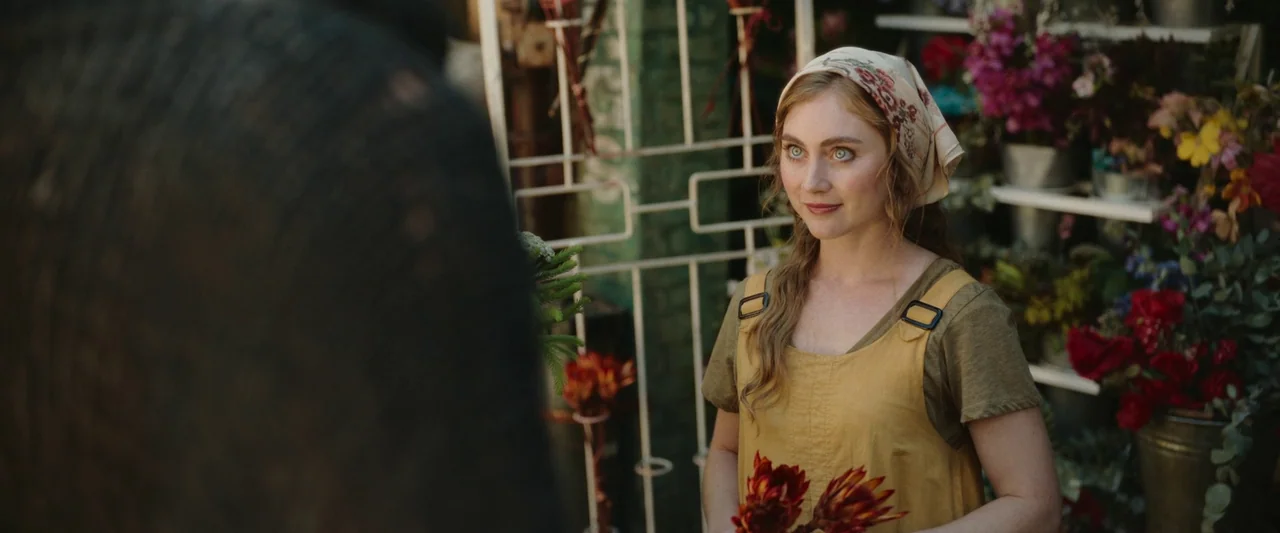 Boy Kills World (2023) movie still: medium — A woman stands in a market stall surrounded by various flower buckets and racks. She holds a cluster of…