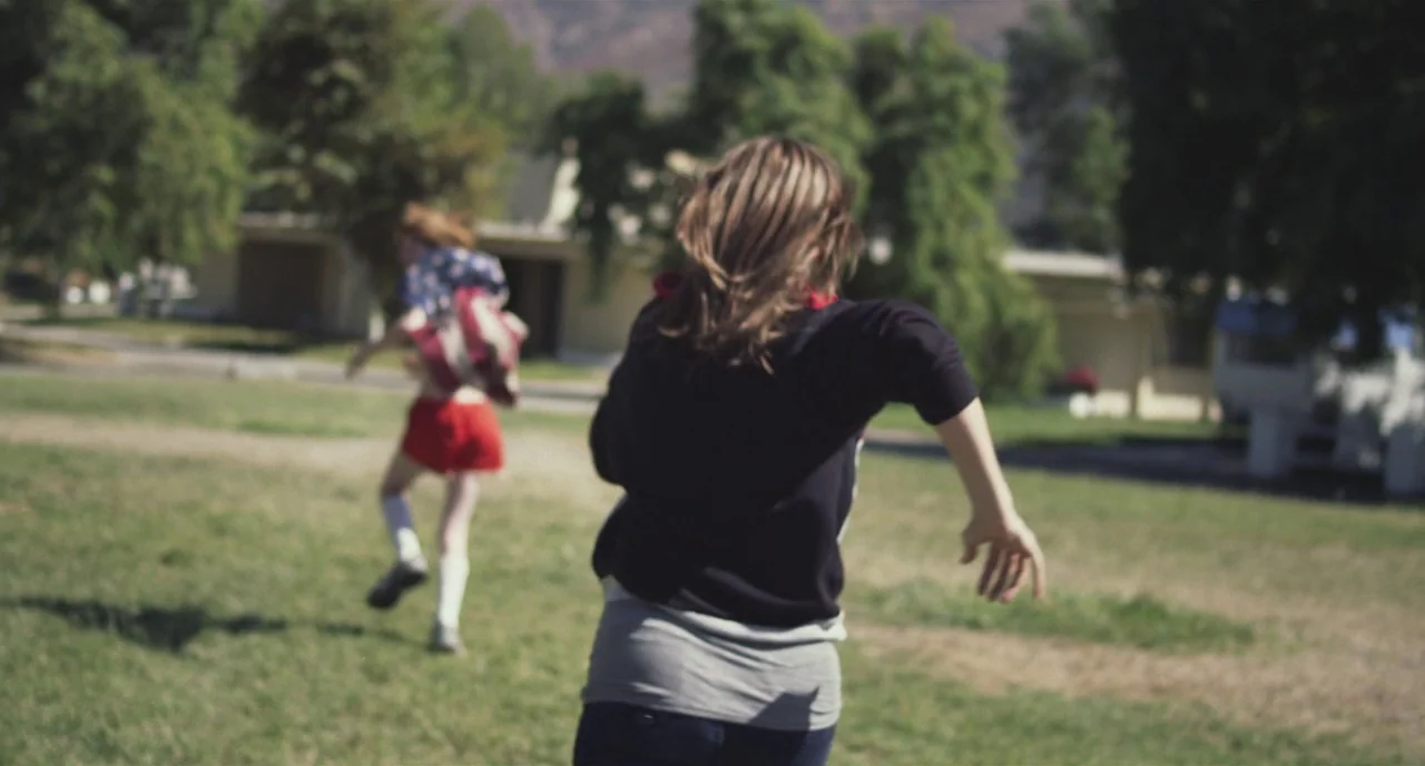 Short Term 12 (2013) movie still: medium — A grassy public park area features mature trees and buildings in the background. Two individuals run…