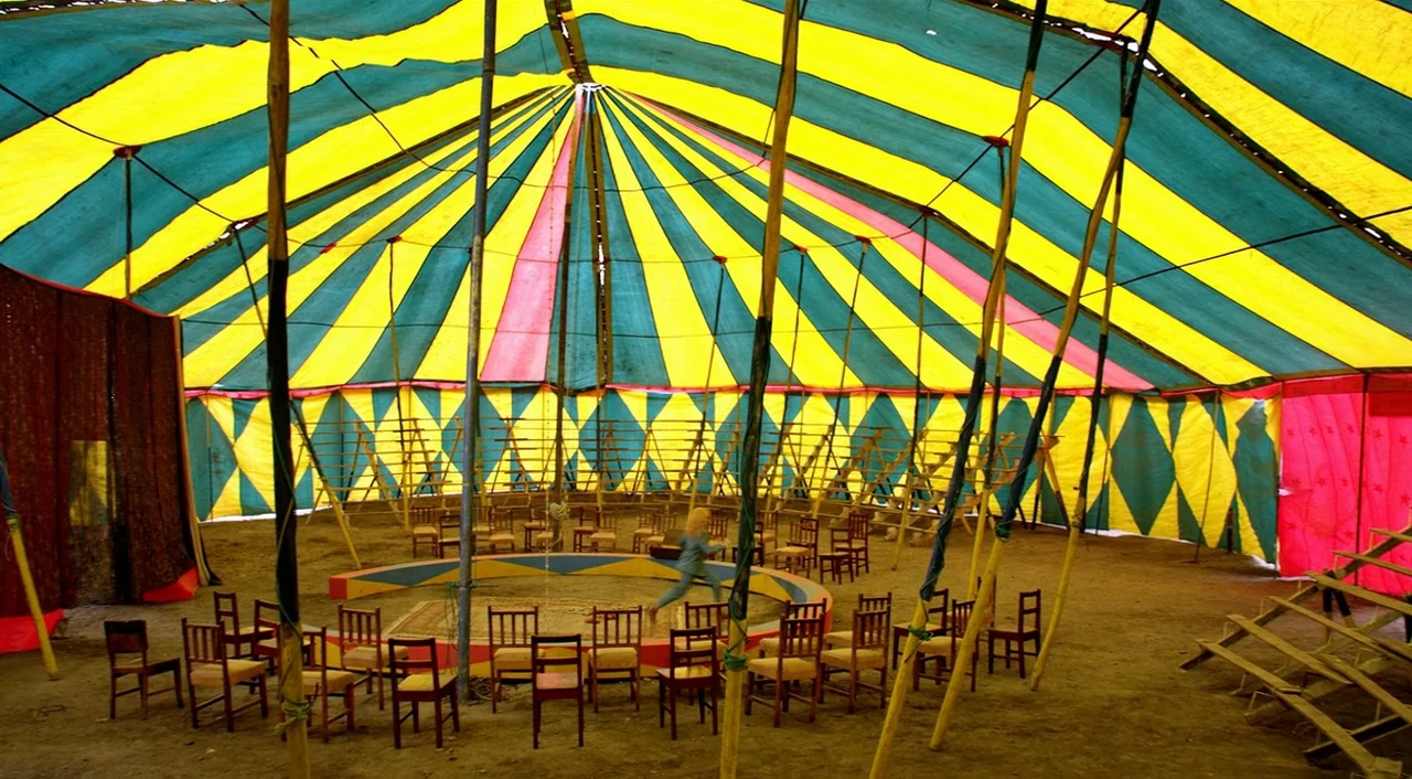 Dance of Reality (2013) movie still: wide — A circus tent interior features a yellow and teal striped ceiling with wooden poles. A small child moves…