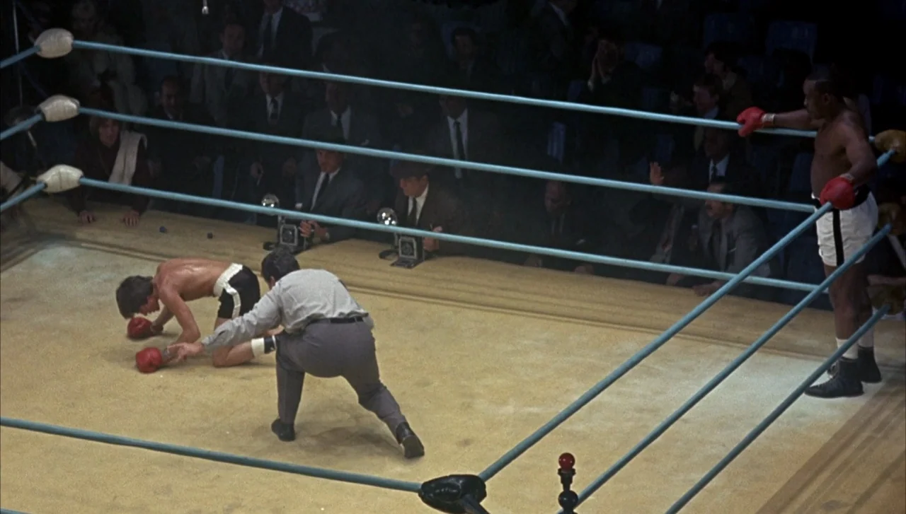 Head (1968) movie still: wide — A brightly lit indoor arena centers on a canvas mat surrounded by blue ropes. A boxer kneels on the floor as a…