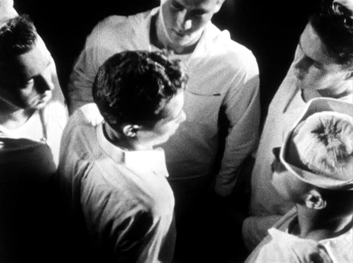 Fireworks (1947) movie still: medium — Four men in white shirts stand gathered together in a dark, enclosed space. They look downward in a circle…