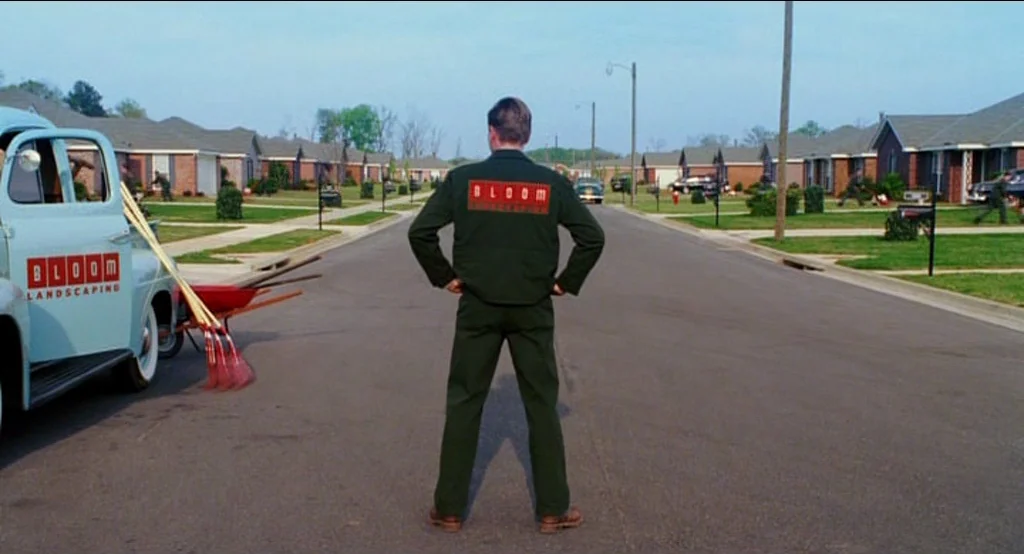 Big Fish (2003) movie still: fullbody — A suburban street lined with uniform single-story houses and manicured green grass lawns. A man in a work…