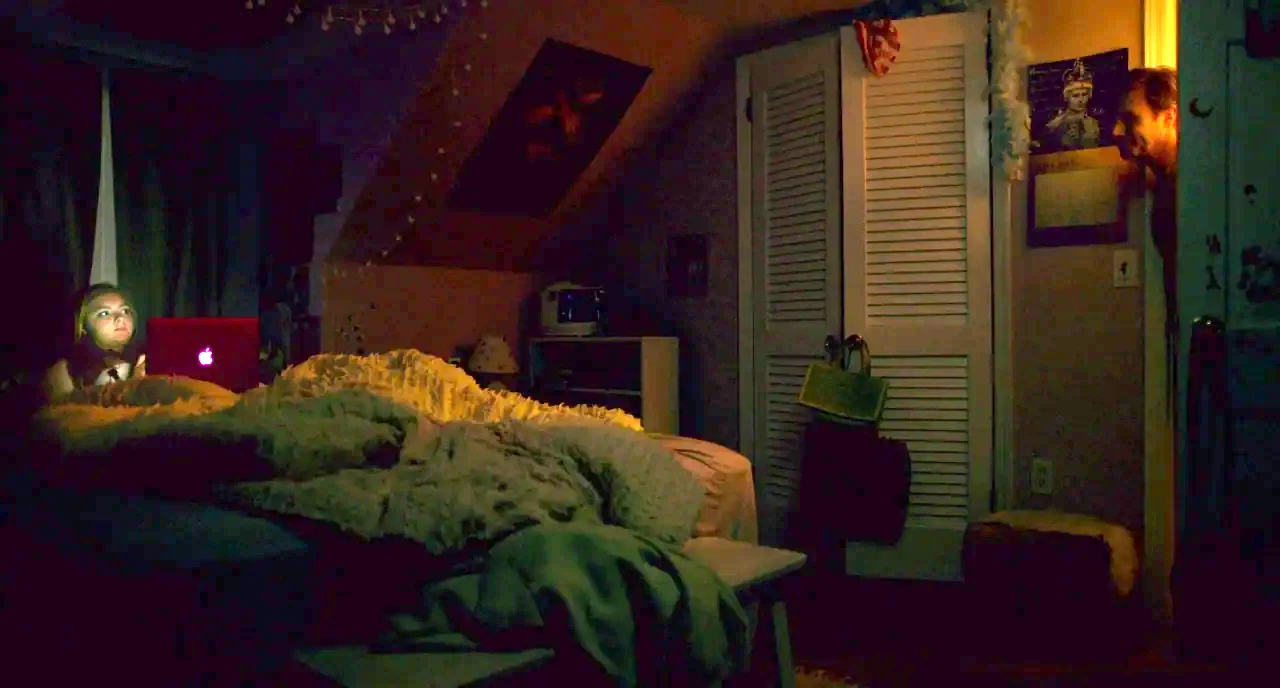 Eighth Grade (2018) movie still: wide — A dark bedroom with white louvered closet doors and a cluttered bed. A woman sits on the bed with a glowing…