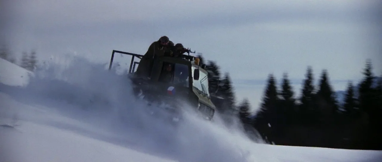 The Living Daylights (1987) movie still: wide — A military vehicle travels through deep snow on a mountain slope with pine trees. Soldiers ride in…