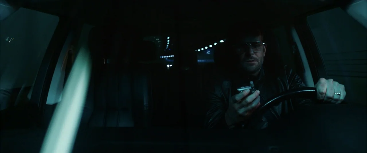 War Dogs (2016) movie still: medium — A dark vehicle interior with the driver visible in the foreground under low light. The man looks down at a…