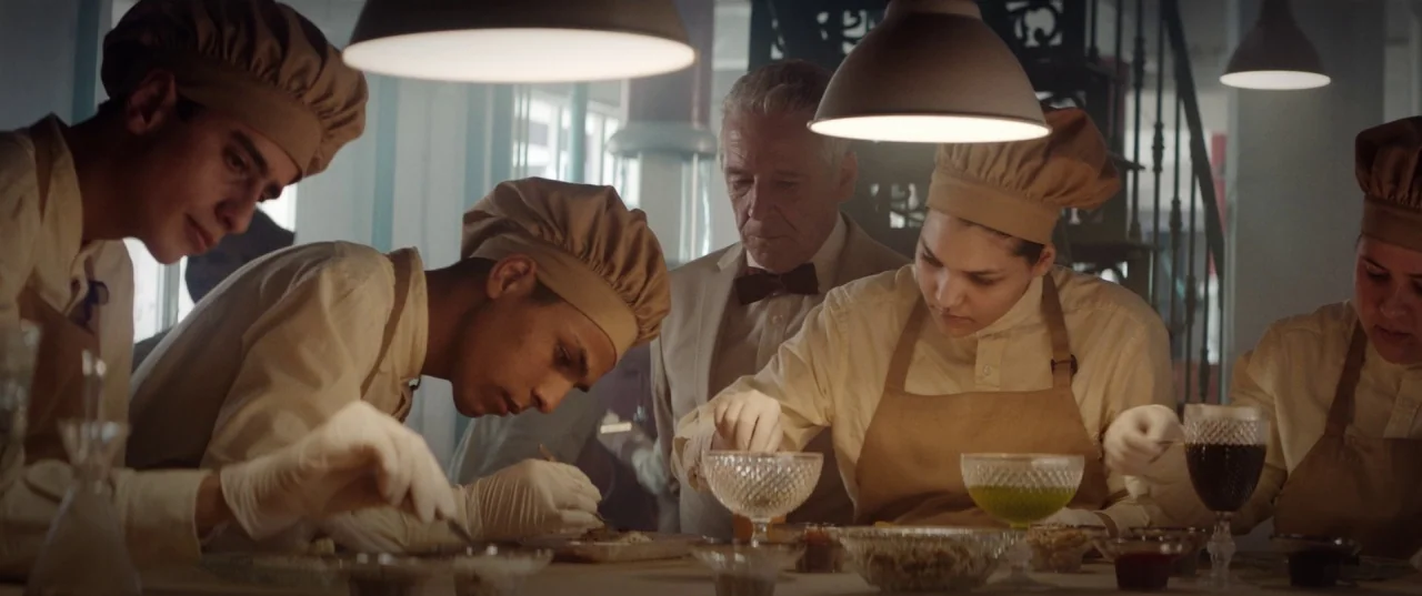 The Platform (2019) movie still: medium — A group of people wearing culinary uniforms work at a long table in a lab. The setting contains various…