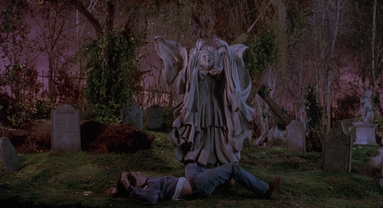 Hocus Pocus (1993) movie still: wide — A dark graveyard at night filled with weathered tombstones and overgrown trees. A man lies motionless on the…