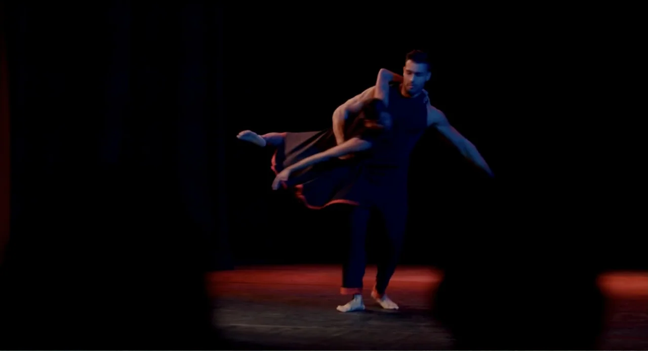 Can You Keep a Secret? (2019) movie still: fullbody — A dark performance space with a wooden floor lit by colored theatrical lights. A man lifts a…