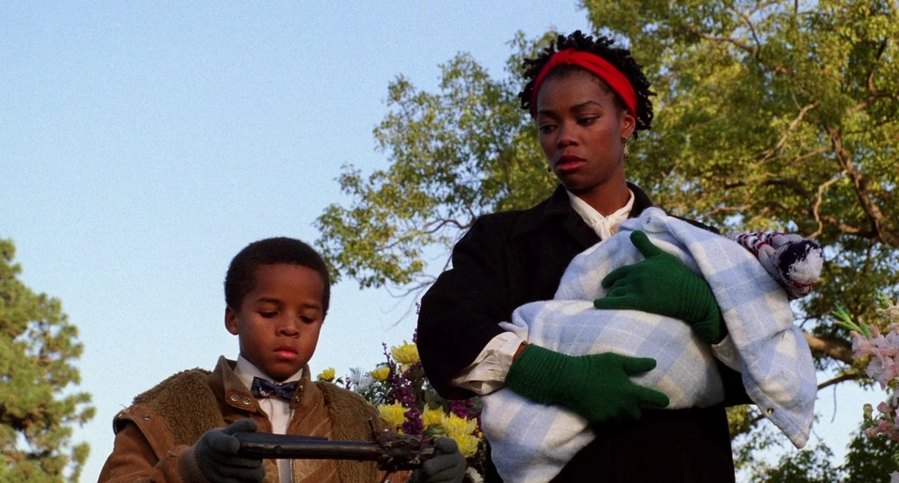 Candyman (1992) movie still: medium — A woman and a young boy stand outdoors against a backdrop of trees. The woman cradles a bundle while the boy…