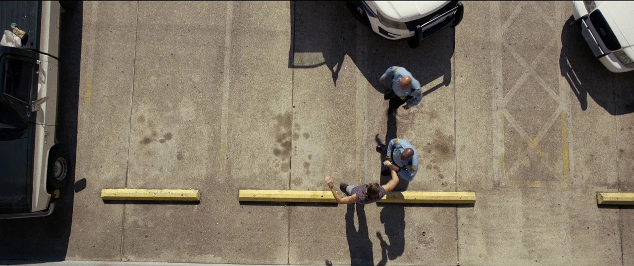 Hit Man (2023) movie still: wide — A concrete parking lot with painted yellow lines and yellow parking bumpers. Two police officers stand over a man…