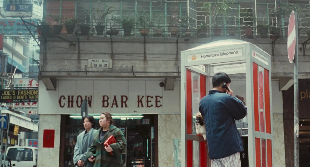 Comrades: Almost A Love Story (1996) movie still: wide — A Hong Kong street features a classic red telephone booth on the sidewalk. Two women walk…