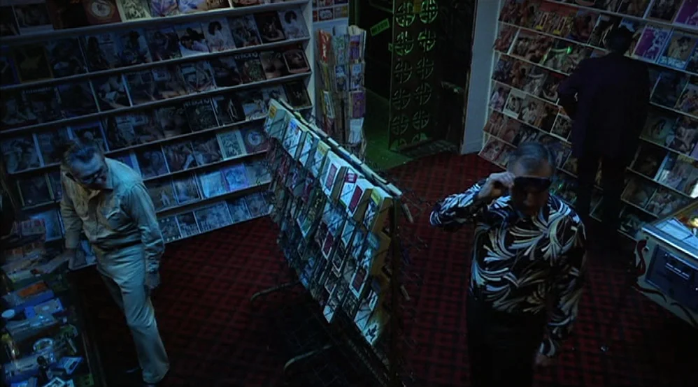 Hardcore (1979) movie still: wide — A dimly lit adult magazine shop features metal racks filled with periodicals and red carpeting. Two men walk…