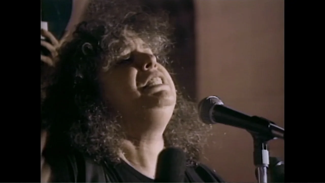 Dear Ms.: A Revolution in Print (2025) movie still: closeup — A dimly lit indoor concert setting with a plain dark background behind the subject. A…