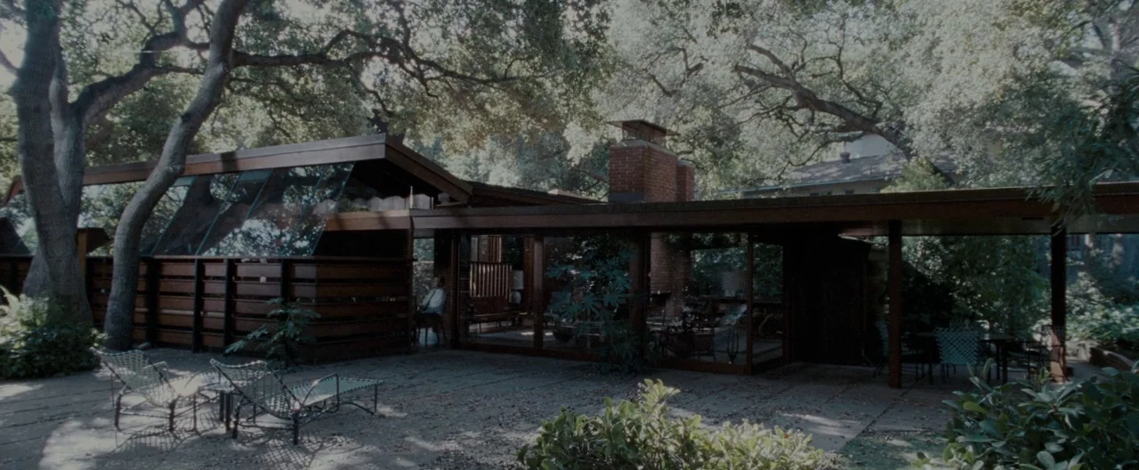 A Single Man (2009) movie still: establishing — A mid-century modern residence with wood siding and large windows sits under tall trees. A brick…