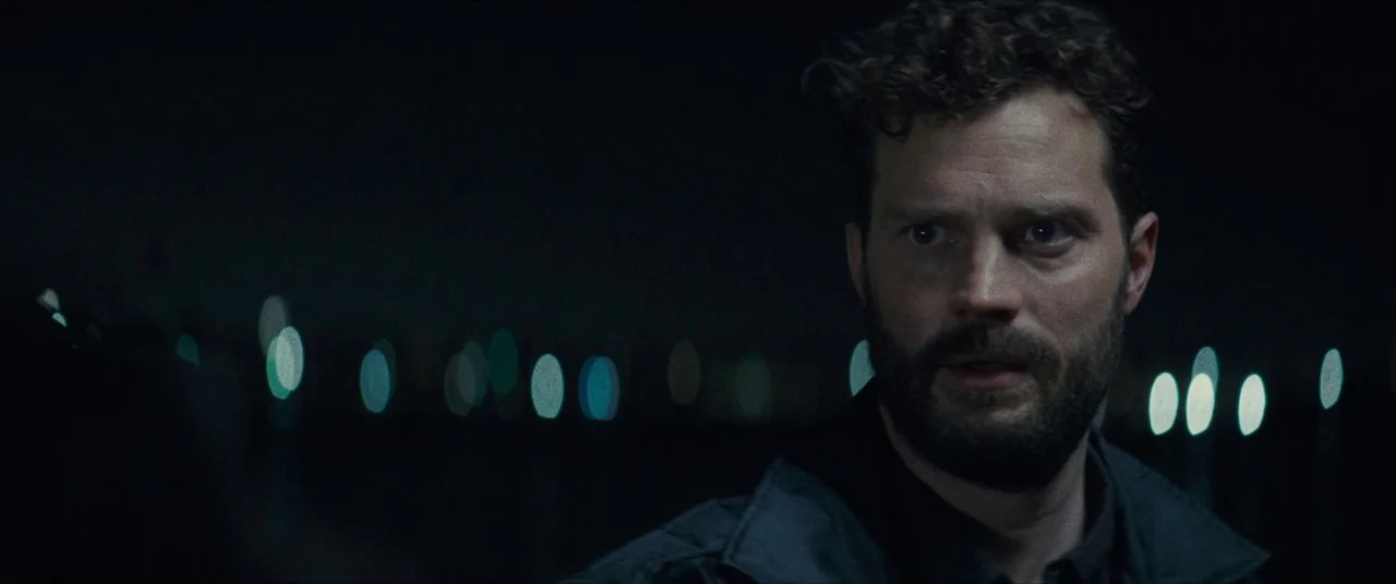 Synchronic (2019) movie still: medium — A man stands outdoors at night near a dark waterfront at night. The background features a horizontal line of…