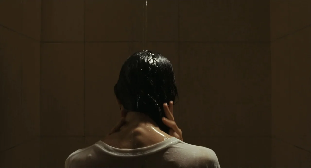 Belén (2025) movie still: medium — A dimly lit shower stall features tiled walls and a single water source overhead. A woman wearing a wet white…