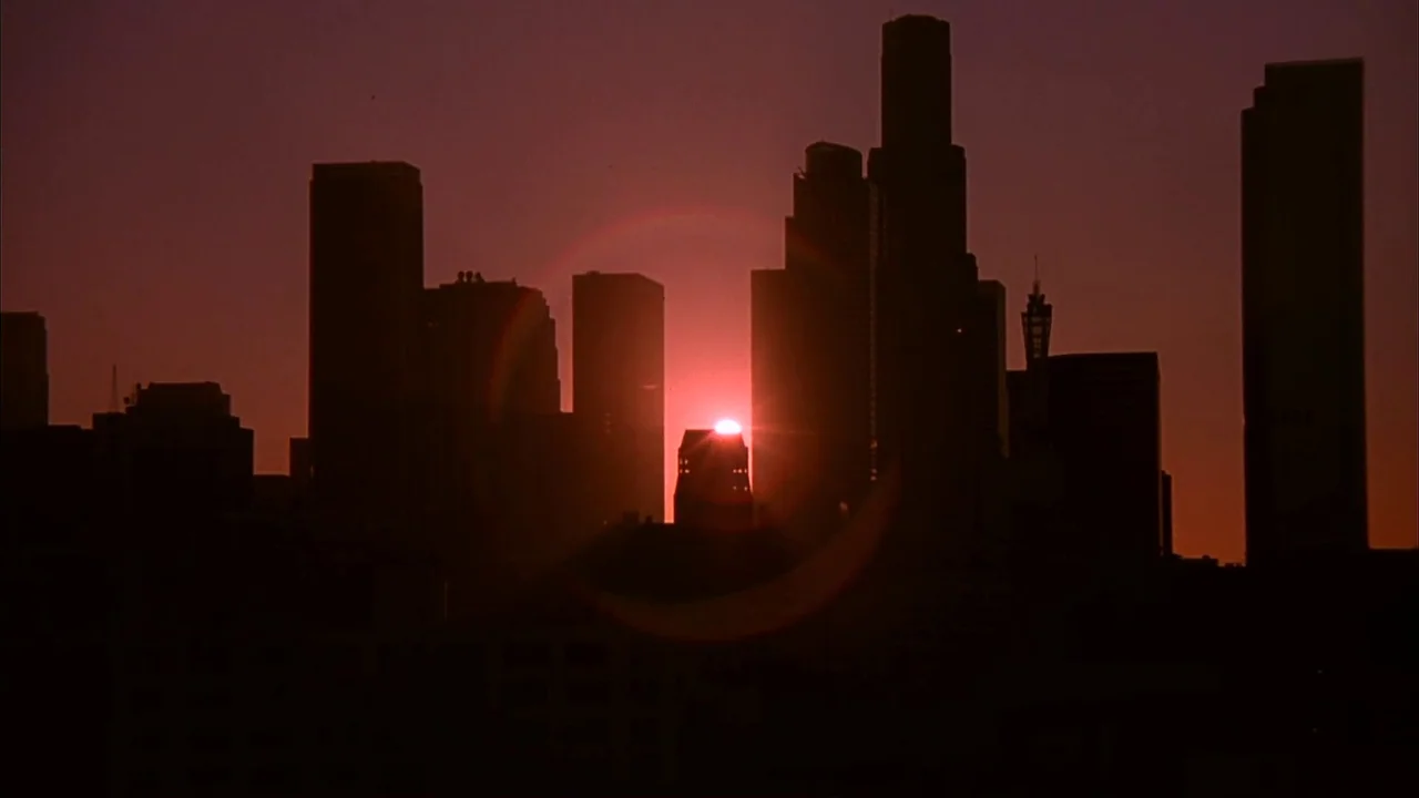 The Mask (1994) movie still: wide — A metropolitan skyline appears as a dark silhouette against a vivid red sunset. The sun is positioned behind a…