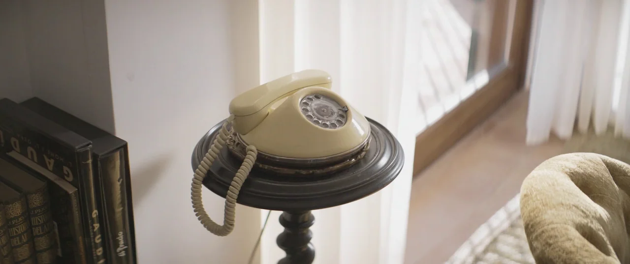 Daaaaaalí! (2023) movie still: closeup — A cream-colored vintage rotary telephone sits atop a dark wooden pedestal near a window. Sunlight filters…