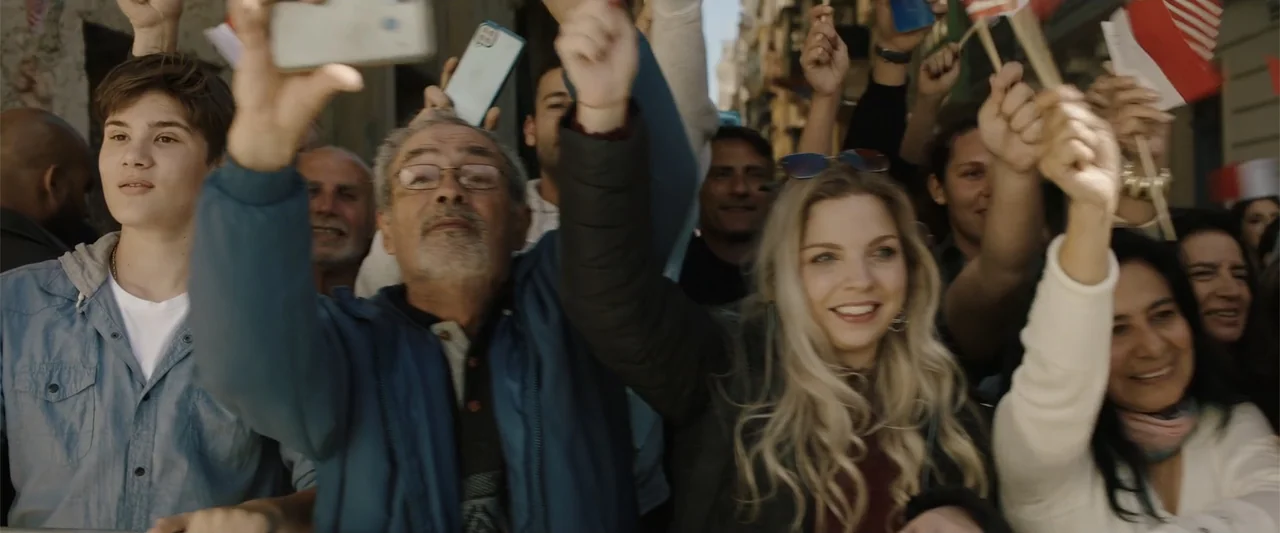 Hounds of War (2024) movie still: medium — A diverse crowd of people gathered outdoors during the day in a busy area. Many individuals hold…