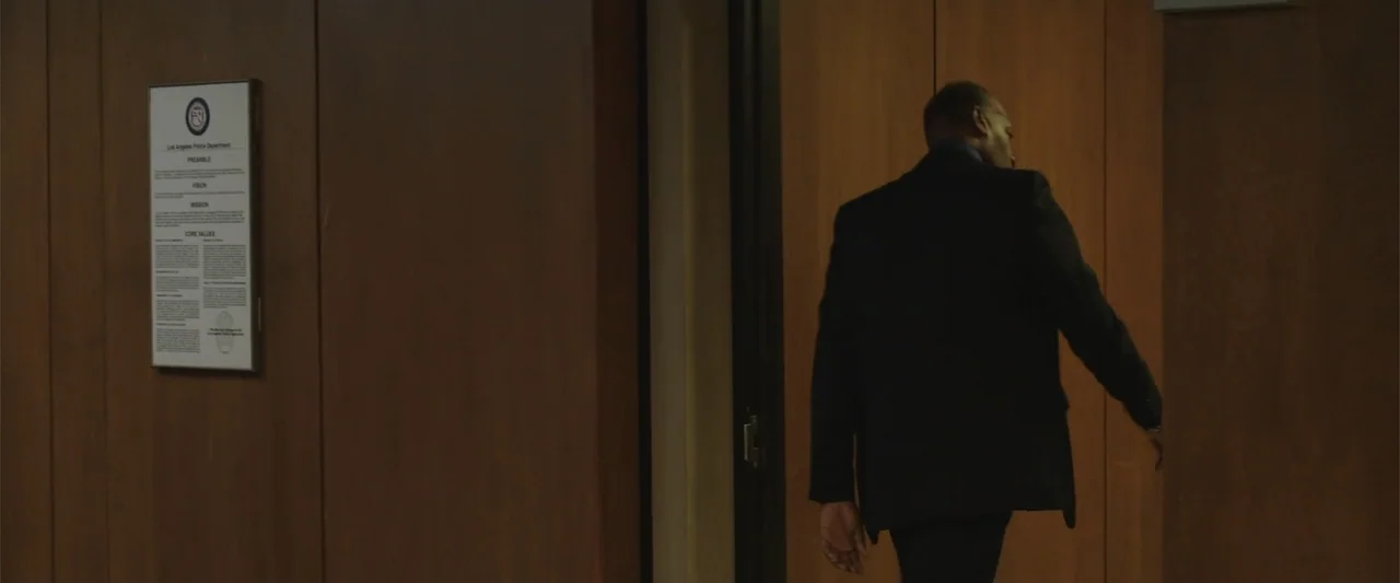 Presagios (2024) movie still: medium — A wood-paneled hallway in a police station featuring a framed mission statement plaque. A man in a suit walks…