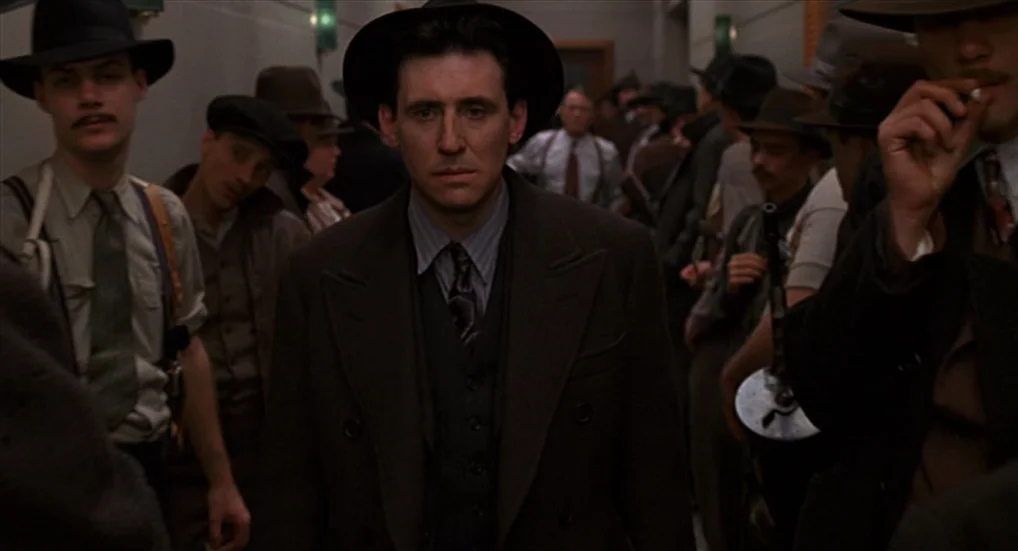 Miller’s Crossing (1990) movie still: medium — A dark hallway filled with men wearing 1930s era fedoras and suits. One man stands in the center…