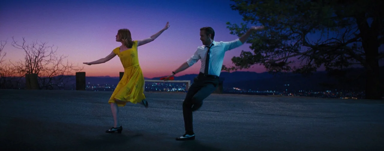 La La Land (2016) movie still: wide — A paved area on a hillside overlooks a city glowing in the dusk light. A man and a woman dance with their arms…