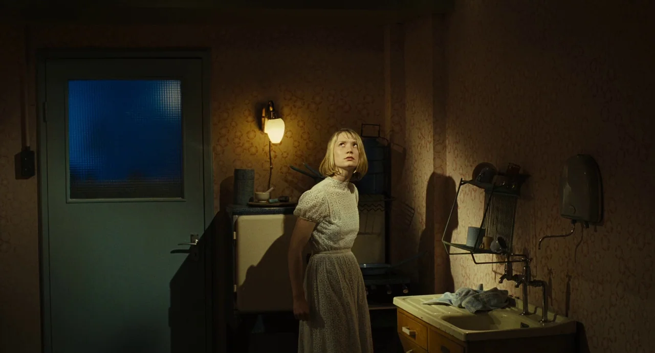 The Double (2013) movie still: medium — A dimly lit room with vintage patterned wallpaper features a sink and a door. A woman stands in the center…