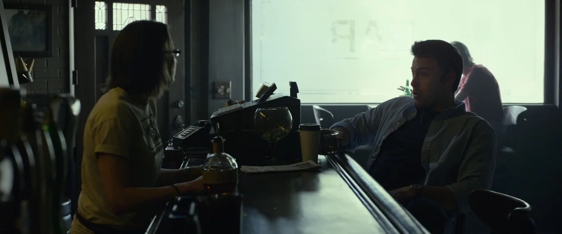 Gone Girl (2014) movie still: medium — A dimly lit room with a dark wooden bar counter and large rear window. A man and a woman sit at opposite ends…