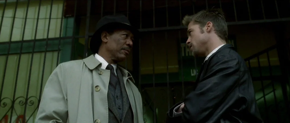 Se7en (1995) movie still: medium — Two men stand in front of a dark metal gate outside a building exterior. The older man in a hat faces the younger…