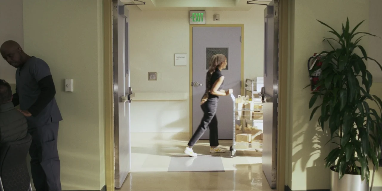 Sitting in Bars with Cake (2023) movie still: fullbody — A clinical hospital hallway features pale walls and a light-colored floor. A woman walks…