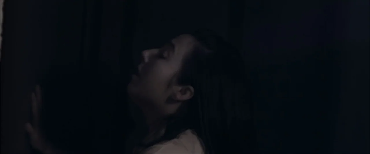 Anomaly (2006) movie still: closeup — A dark, dimly lit interior space with a plain wall as the backdrop. A woman stands in profile against the wall…