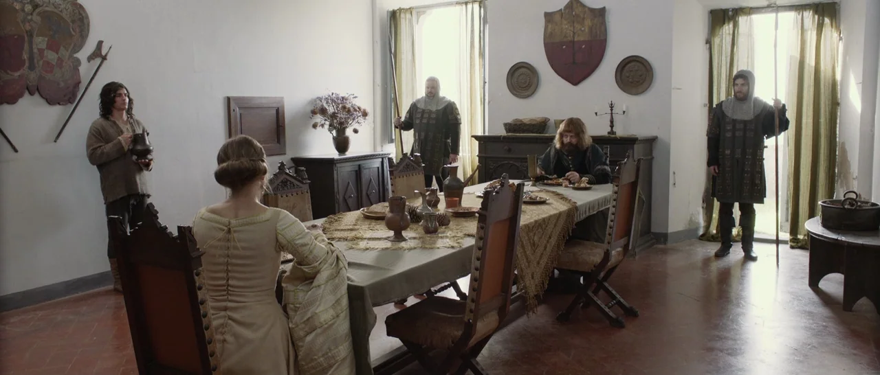 The Little Hours (2017) movie still: wide — A medieval dining room features stone walls, wooden furniture, and decorative heraldic shields…