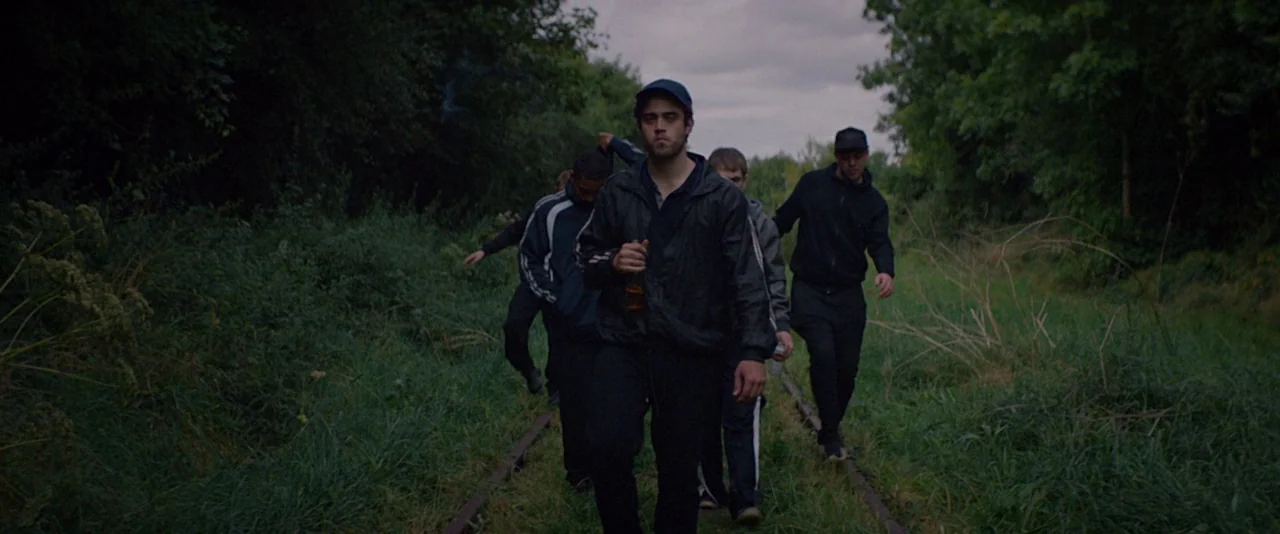 Kissing Candice (2017) movie still: fullbody — A group of men walks along abandoned train tracks surrounded by dense, green vegetation. The overcast…