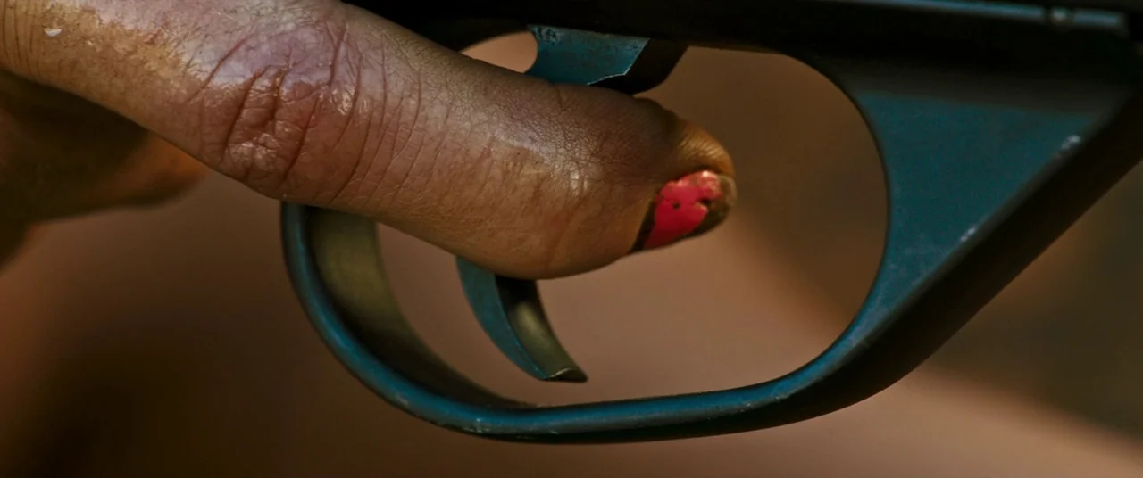 Revenge (2017) movie still: closeup — A close-up view of a finger resting against the trigger of a metal firearm. The fingernail is painted with…
