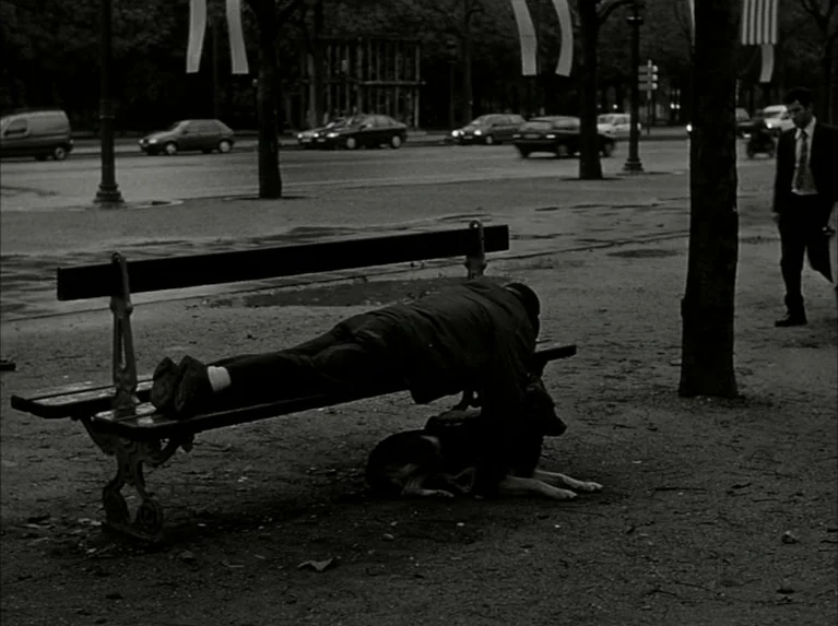 Eloge de L’amour (2001) movie still: wide — A public park area features a dark wooden bench positioned along a paved road. A person lies prone on…