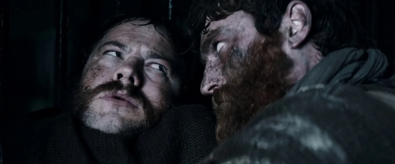 Black ’47 (2018) movie still: closeup — A dimly lit, claustrophobic stone enclosure with vertical iron bars in the background. Two bearded men…
